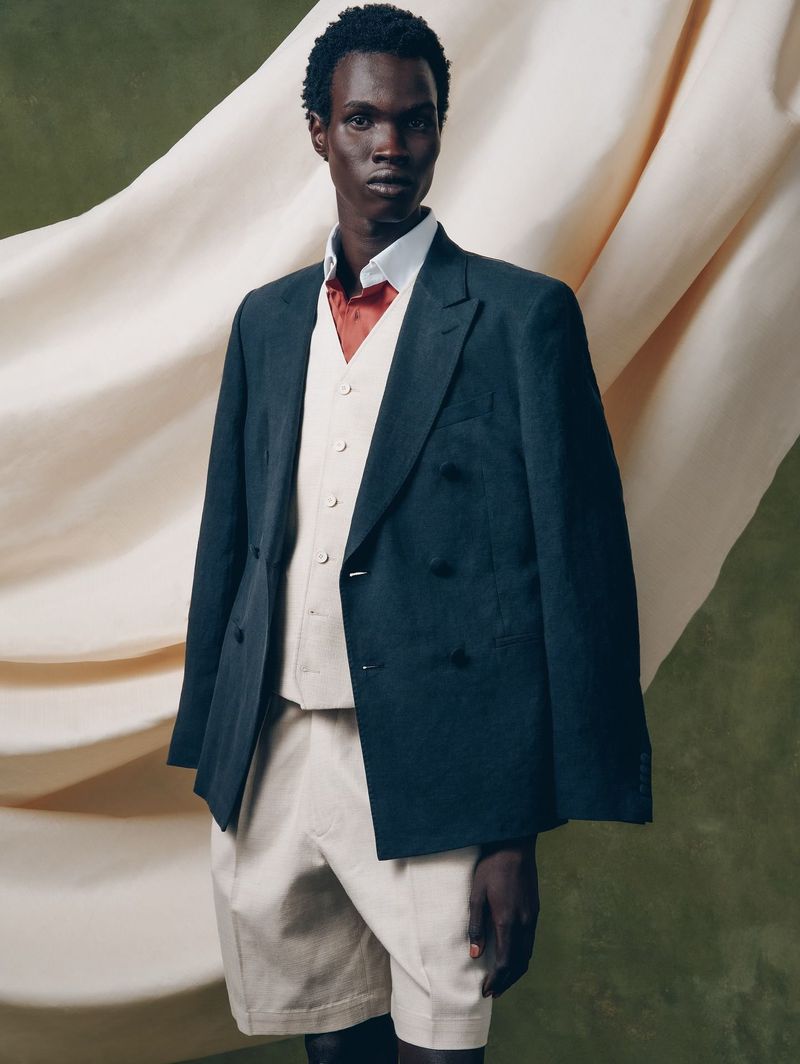 A dark-skinned male fashion model poses indoors wearing a blue blazer, tan vest, white shirt and beige shorts against draped fabric.