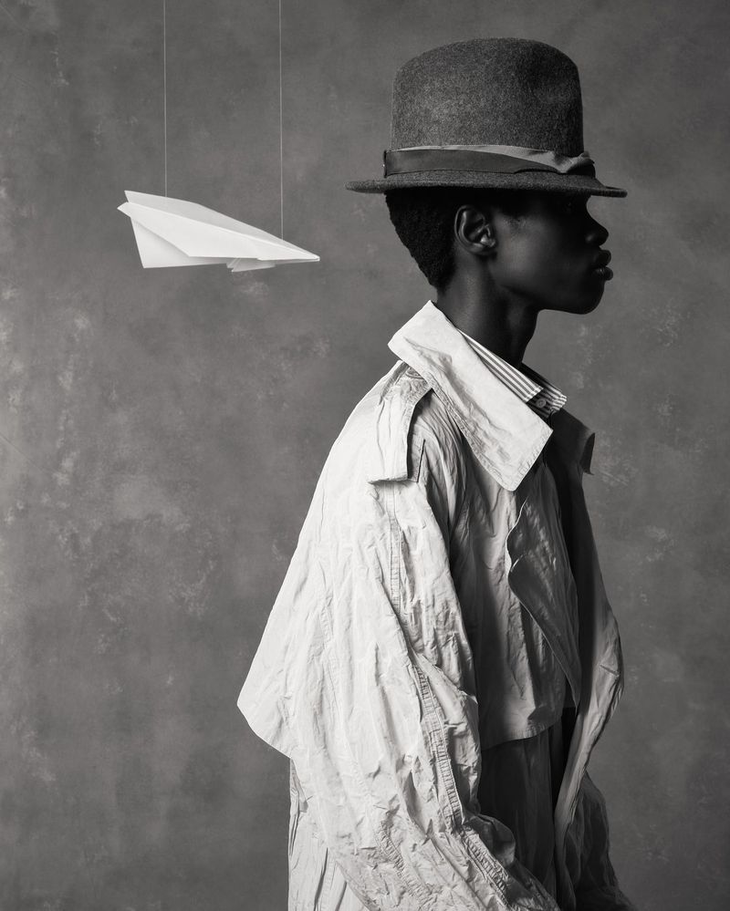 Monochrome portrait of an African man wearing a hat and trench coat in profile, with a paper airplane hanging in the background.