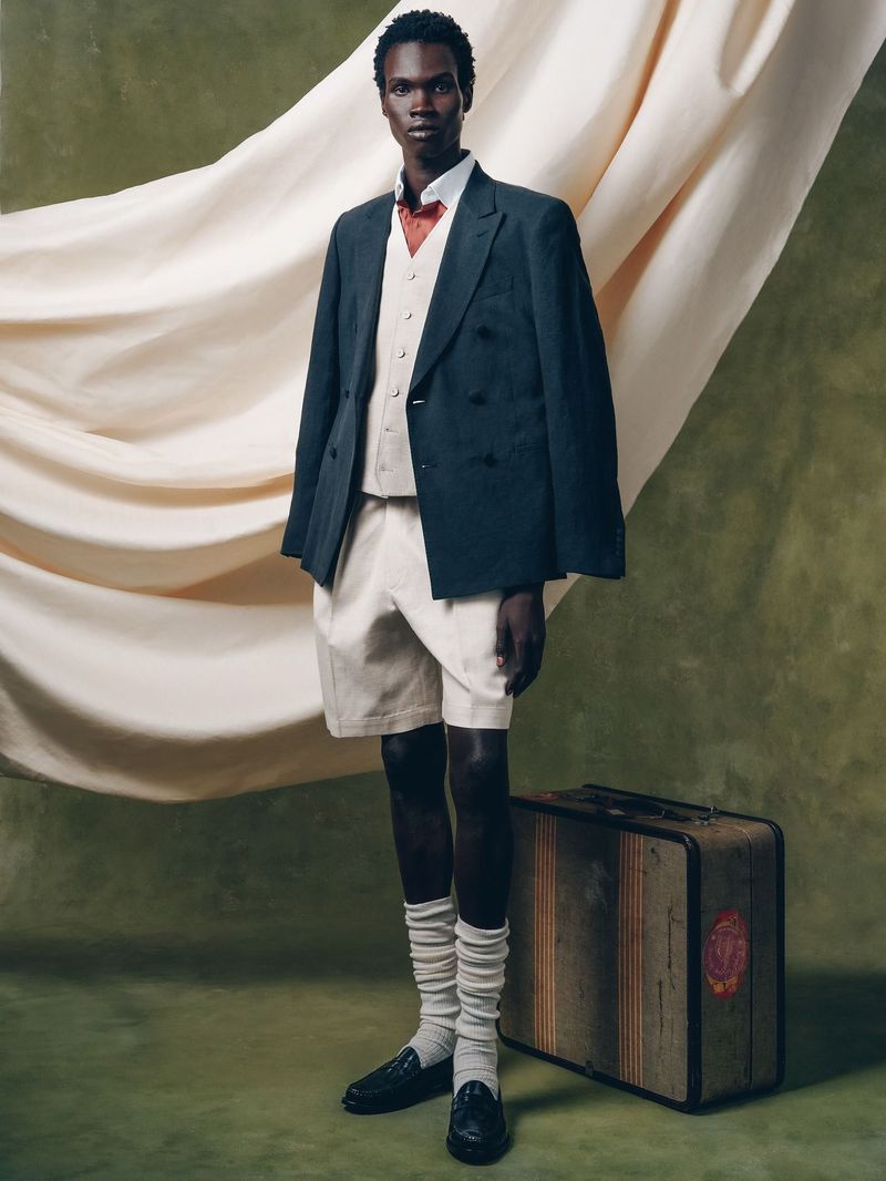 An adult male fashion model poses in a blazer, vest, shorts, and knee-high socks next to a vintage suitcase against a draped backdrop.