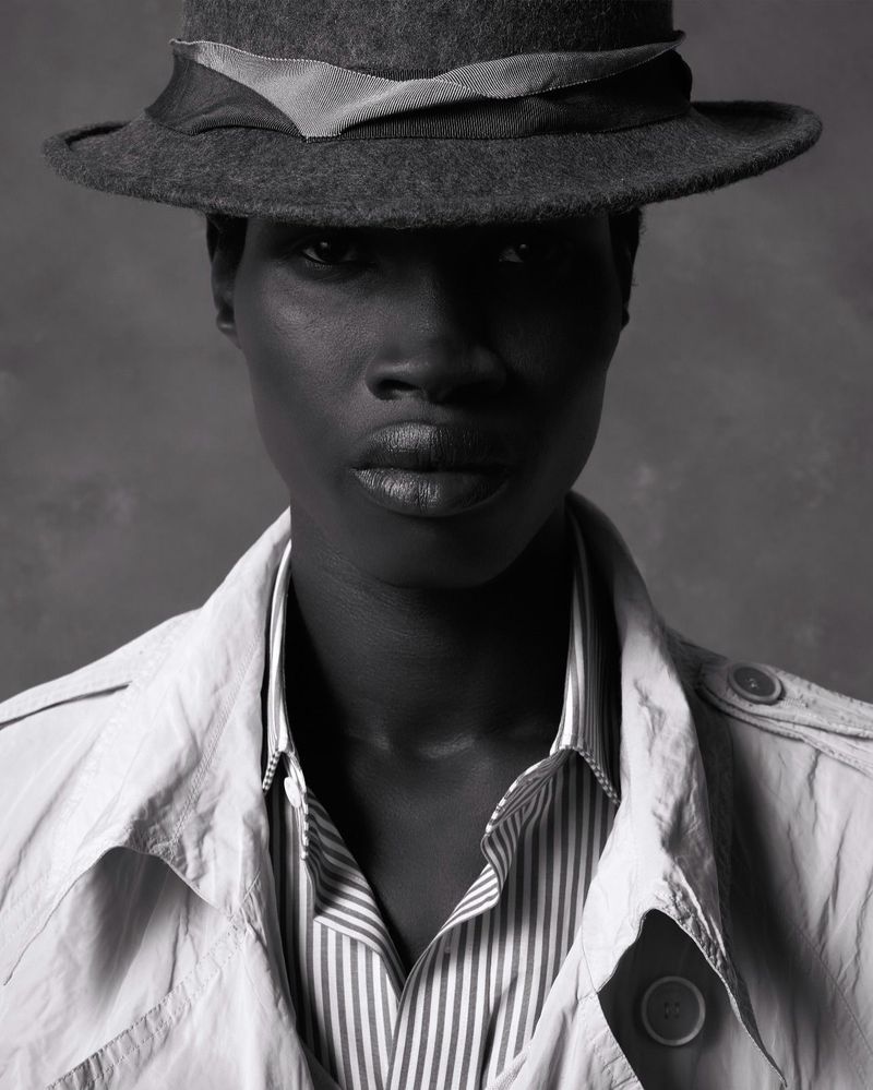 Striking black and white portrait of a dark-skinned woman wearing a hat, trench coat, and striped shirt against a grey backdrop.