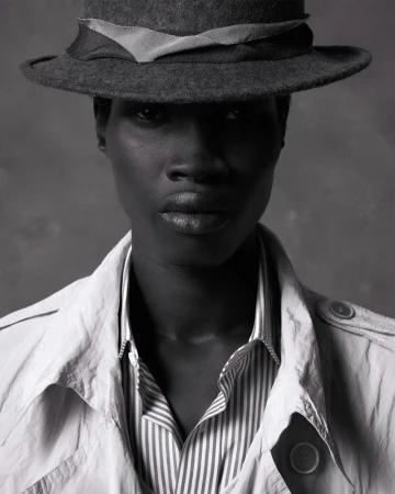 Striking black and white portrait of a dark-skinned woman wearing a hat, trench coat, and striped shirt against a grey backdrop.