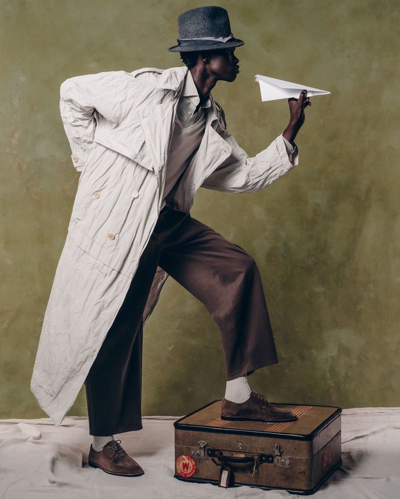 A Black male model with a gray hat and trench coat stands on a vintage suitcase holding a paper airplane in a fashion editorial setting.