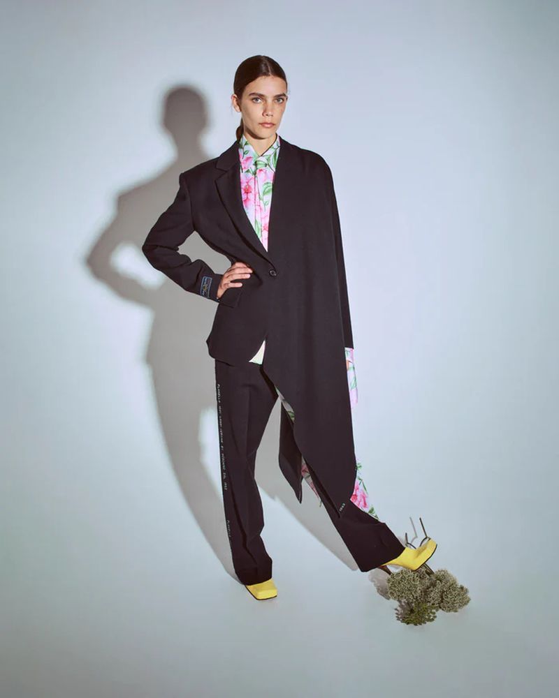 A woman poses in a black blazer, floral blouse, and black pants, standing on bouquet in a bright studio for a fashion editorial.