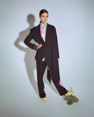 A woman poses in a black blazer, floral blouse, and black pants, standing on bouquet in a bright studio for a fashion editorial.