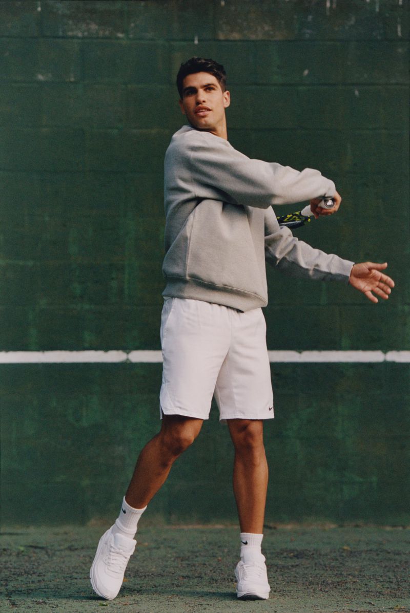 Male tennis player in gray sweatshirt and white shorts prepares to hit a forehand on a green outdoor court with a white line.