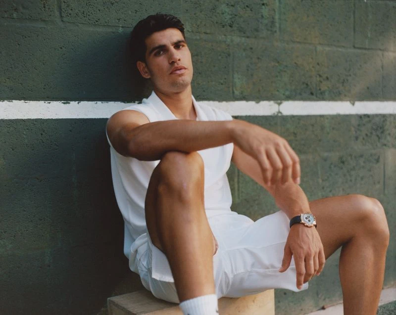 Man in white athletic wear poses against a textured green wall, sitting on a concrete block, showcasing a wristwatch in soft daylight.
