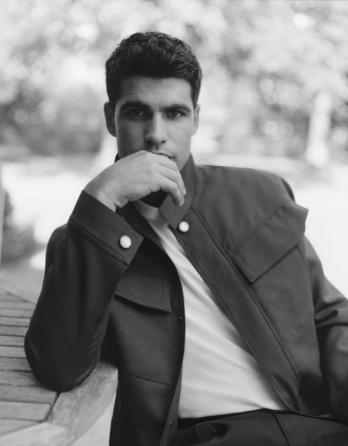 Monochrome portrait of adult male outdoors wearing jacket, sitting on a bench with a blurred leafy background, gazing directly at camera.