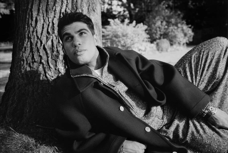 Monochrome outdoor fashion portrait of a man reclining against a tree trunk, wearing an overcoat and tweed pants, gazing upward.