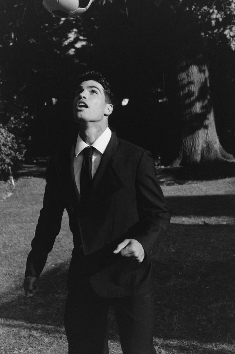 Carlos Alcaraz in a tailored black suit and tie looking up at a soccer ball in an outdoor park setting.