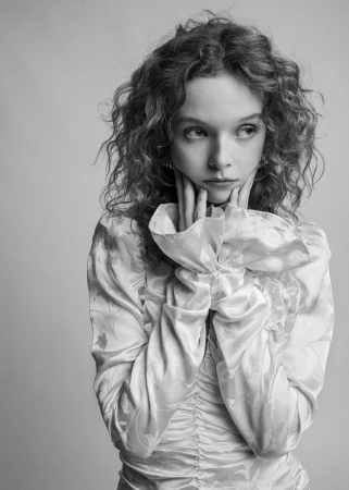 Portrait of a young woman with curly hair in a floral dress, holding her face in her hands against a neutral background in black and white.