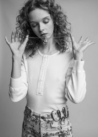 Black and white studio portrait of a young woman with curly hair wearing a white shirt and floral pants, posing with necklace.