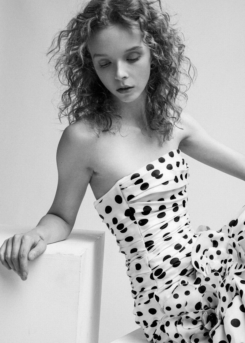 Young woman in a polka dot dress poses in a studio setting. The black and white image showcases the model's curly hair and the dress's pattern.