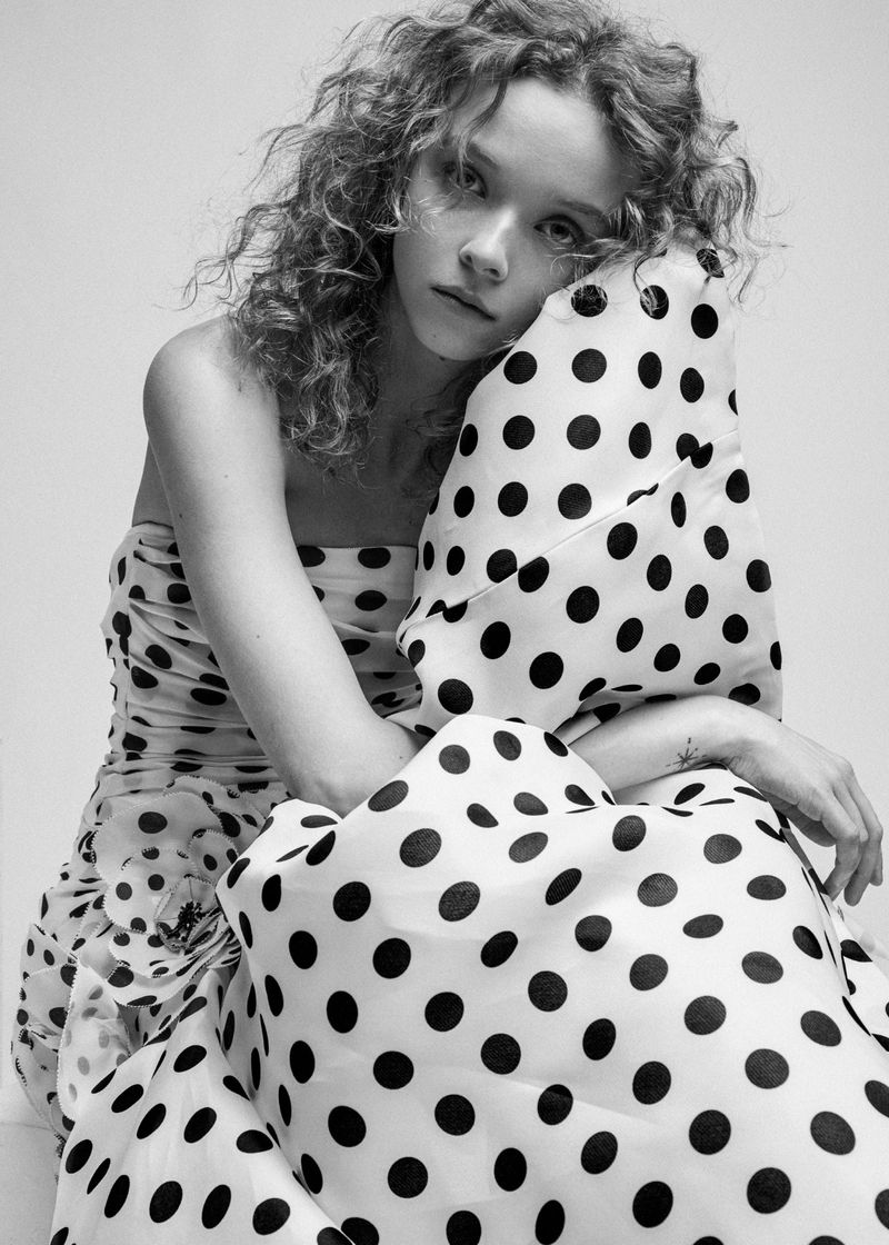 Black and white portrait of a woman in a polka dot dress with curly hair, posing in front of a neutral background in a studio.