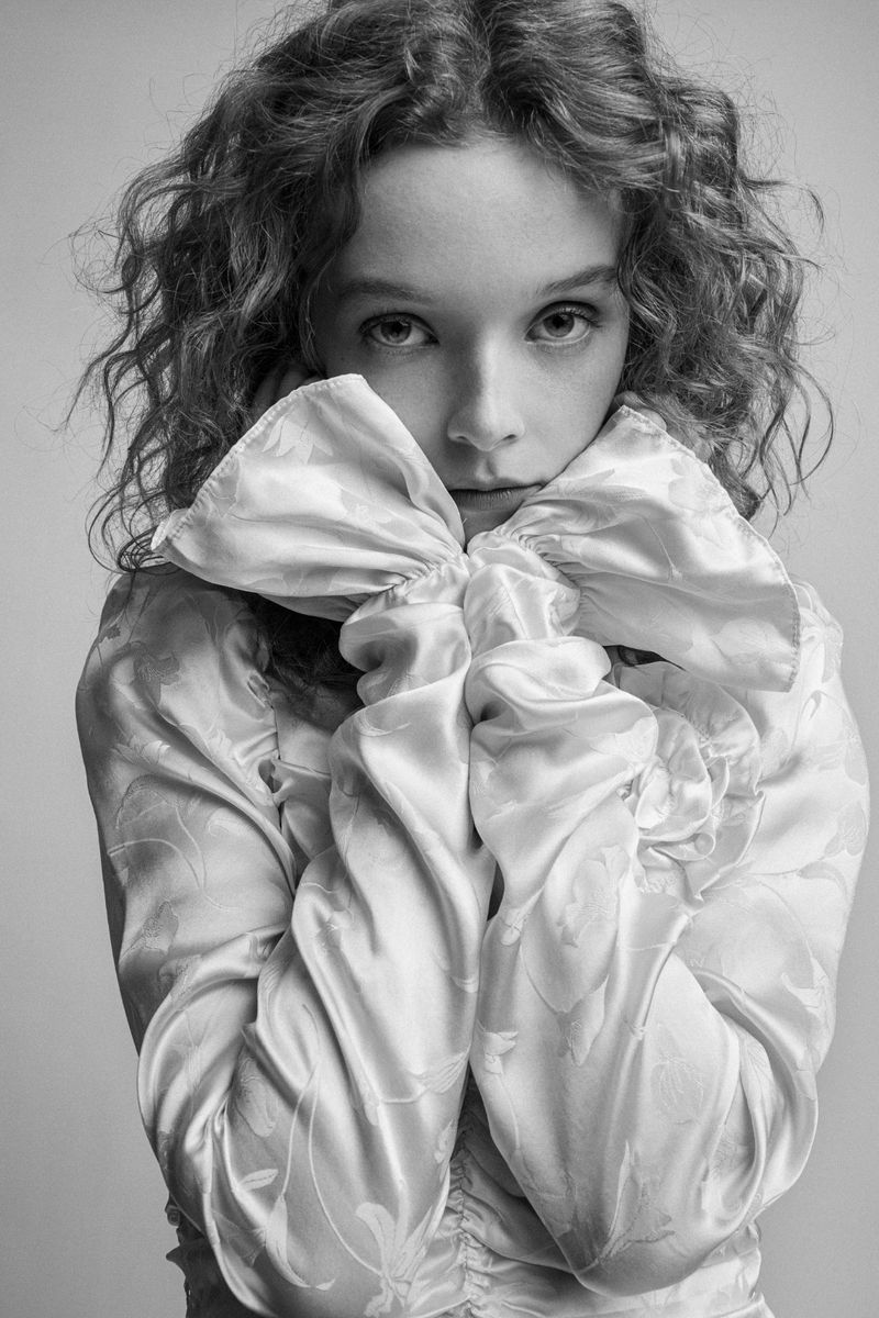 Black and white portrait of a young woman with curly hair, wearing a satin top with large bow, looking directly at the camera.