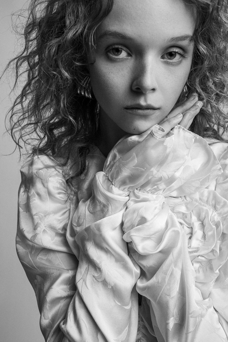 Black and white portrait of model Lily Nova in a ruffled floral jacquard top with hands resting near her chin.