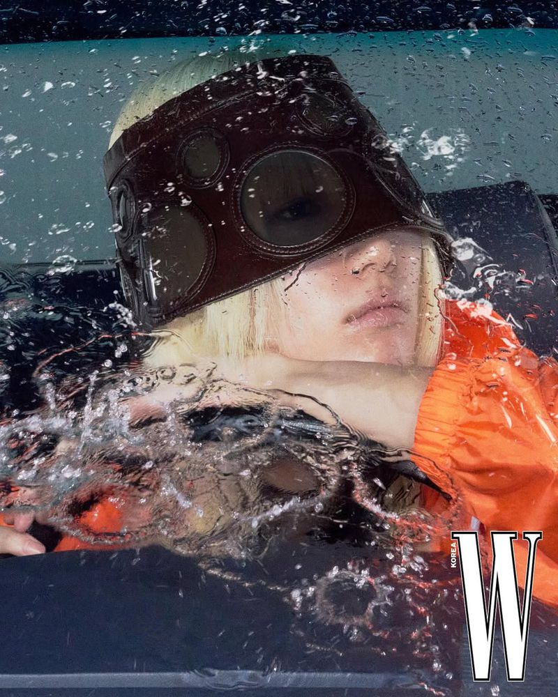 Fashion shot of a fair-skinned person wearing a brown eye covering, with water splashing and an orange sleeve visible in a car.
