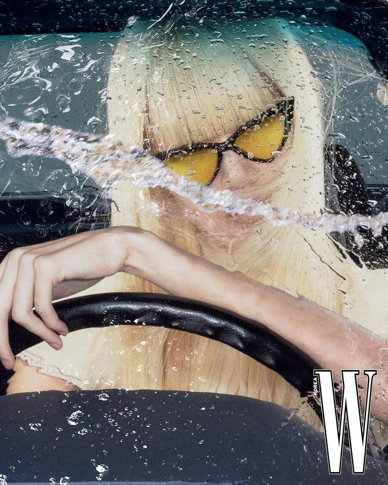 Woman with blonde hair, sunglasses driving a car in the rain. Close-up of driver with water droplets on windshield and car window.