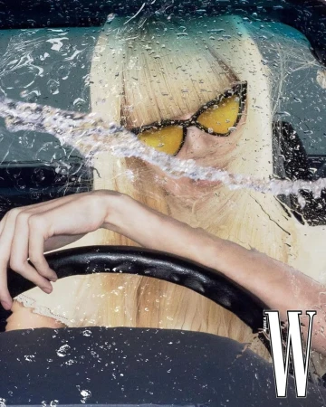 Woman with blonde hair, sunglasses driving a car in the rain. Close-up of driver with water droplets on windshield and car window.