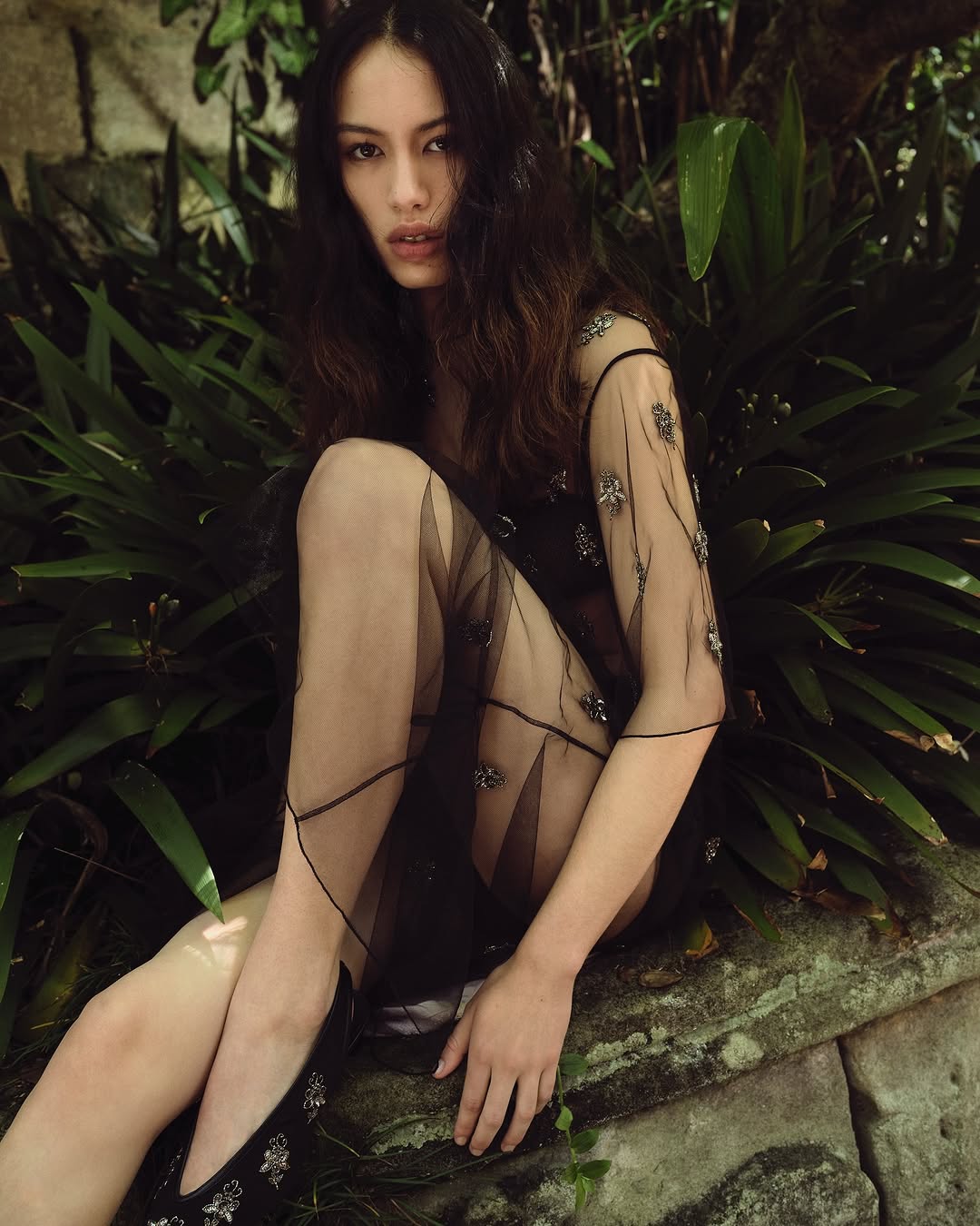 An Asian woman poses in a sheer black dress with floral embellishments, seated on a stone surface with a lush green foliage backdrop.