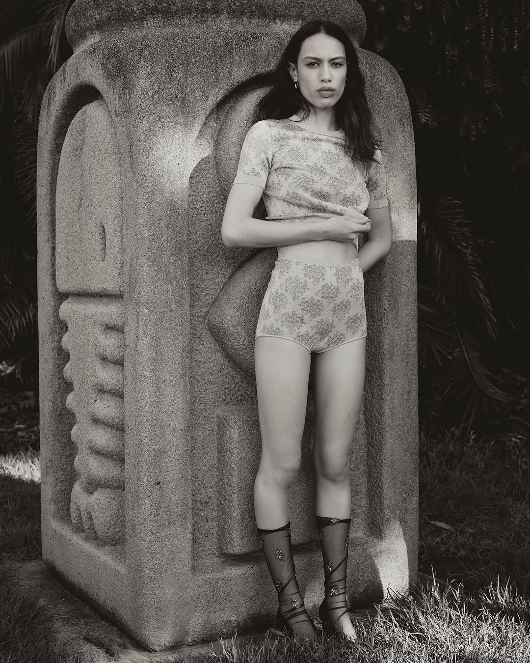A woman in patterned underwear and top stands next to a stone carved object outdoors. The image is in black and white.