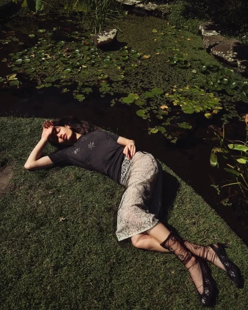 Woman in sheer socks and floral top lies on green grass in a garden near pond with lily pads, bathed in sunlight during daytime.