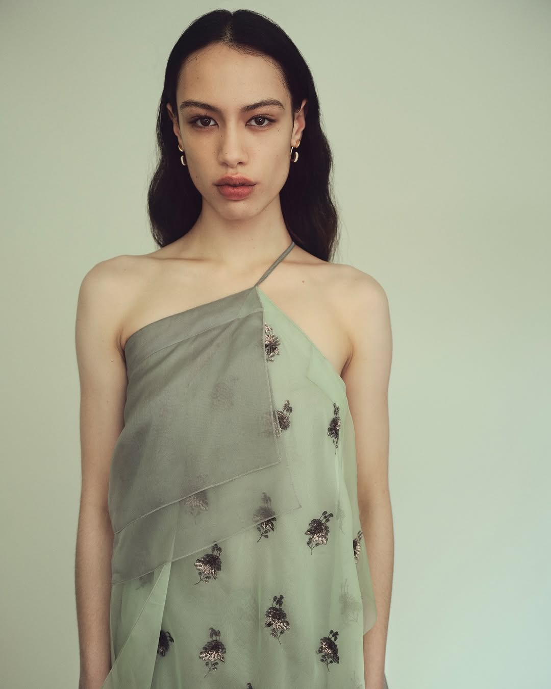 A slender young woman with dark hair poses in a light green and grey one-shoulder floral-patterned dress against a neutral background.