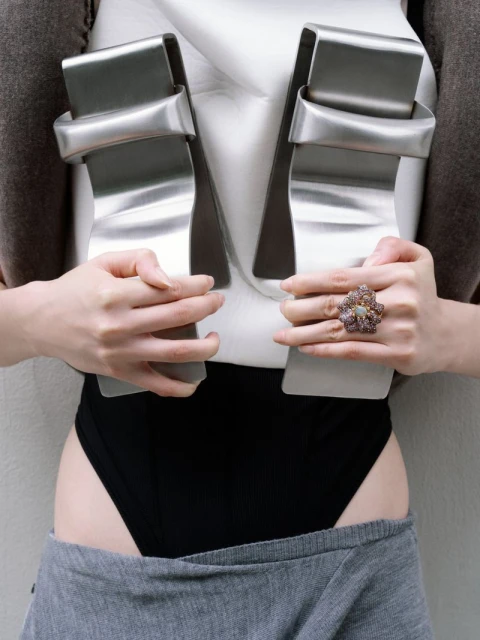 Fashionable studio shot of a model displaying silver sculptural clothing with a large ring. Silver, white, gray, and black tones dominate.