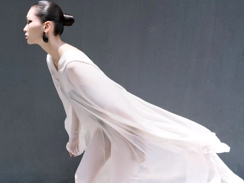 Woman in flowing white dress poses in profile against grey background, accessorized with dark teardrop earrings and a sleek updo hairstyle.
