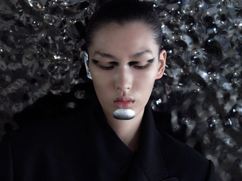 Close-up portrait of a model with silver makeup and black eye shadow against a metallic textured background wearing a black top.