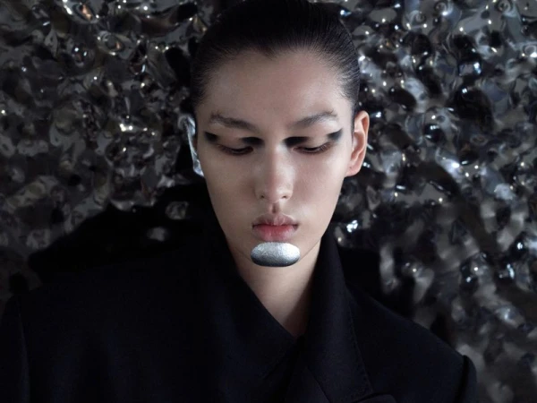 Close-up portrait of a model with silver makeup and black eye shadow against a metallic textured background wearing a black top.