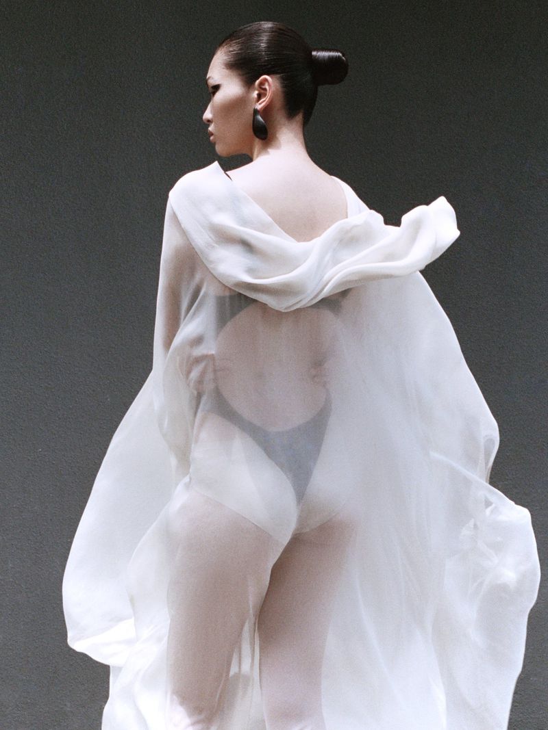 Fashion shot of an adult female posing with her back to the camera wearing a flowing white garment in front of a grey background.