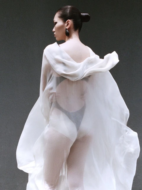 Fashion shot of an adult female posing with her back to the camera wearing a flowing white garment in front of a grey background.