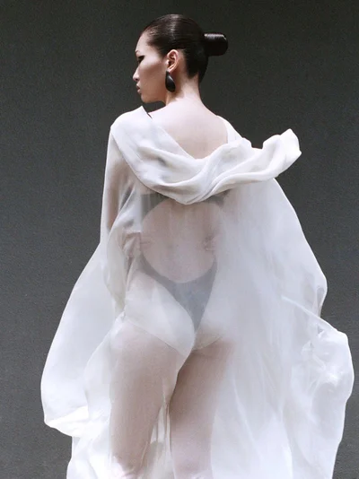 Model Ying Ouyang viewed from behind wearing a billowing translucent white cape and black bodysuit against a dark grey backdrop.