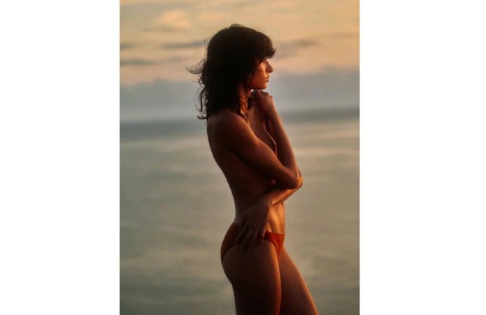 Woman in profile posing against a sky and water background, wearing an orange swimsuit bottom, and looking into the distance.