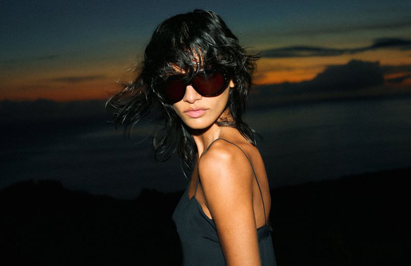 A woman with black hair and sunglasses poses in a black top against a dark horizon with an orange-tinted sky at twilight.
