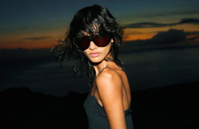 A woman with black hair and sunglasses poses in a black top against a dark horizon with an orange-tinted sky at twilight.
