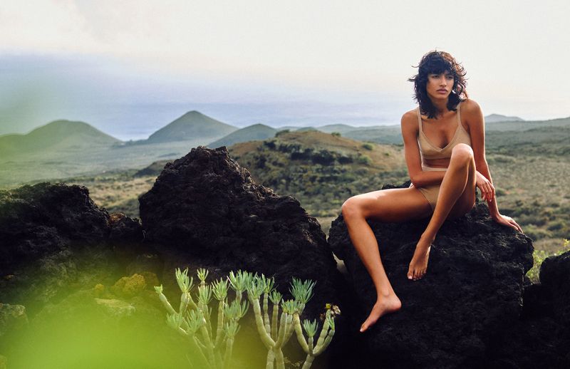 A woman in a tan swimsuit sits on a rock formation with legs extended, with mountains visible in the background in outdoor setting.