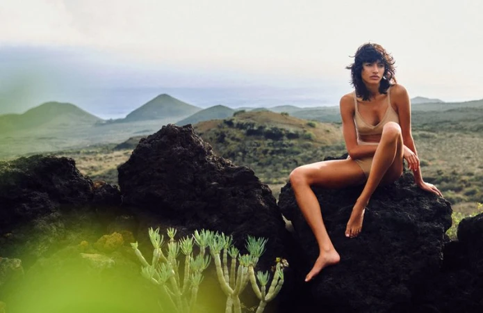 A woman in a tan swimsuit sits on a rock formation with legs extended, with mountains visible in the background in outdoor setting.