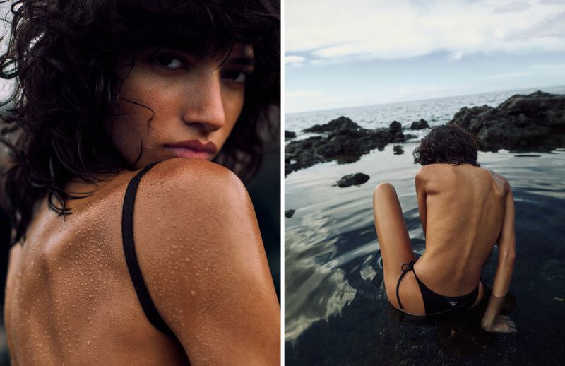 Woman in black swimwear sits in water by a rocky beach with a serene expression, showcasing a sun-kissed back and the sea backdrop.
