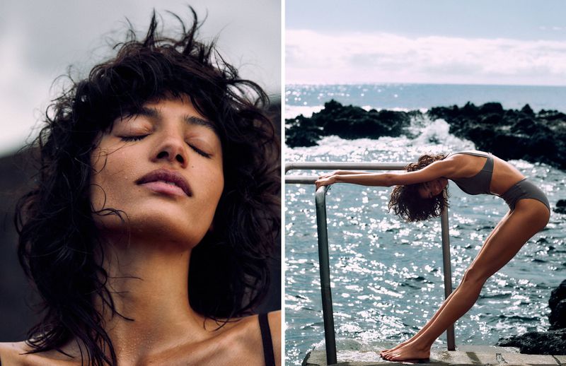 Woman with closed eyes, wet hair poses in sunlight. Woman in swimwear arches back near rocky coast and ocean with sunlight reflection.