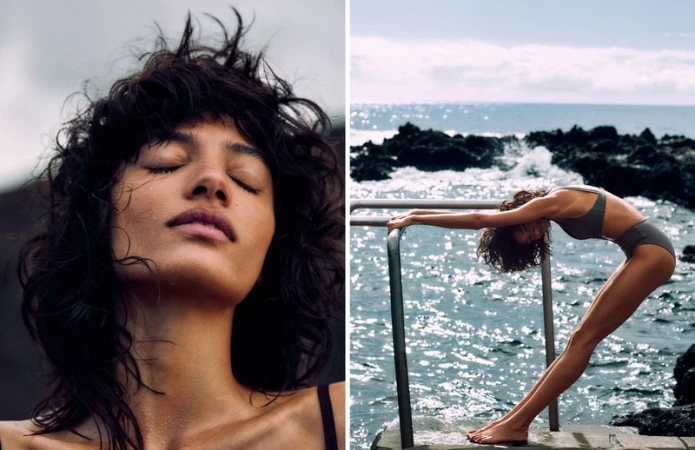 Woman with closed eyes, wet hair poses in sunlight. Woman in swimwear arches back near rocky coast and ocean with sunlight reflection.