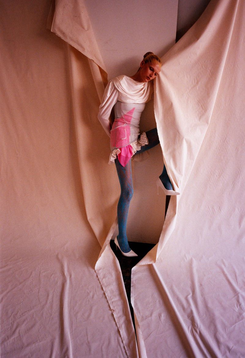 Fashion photo featuring a slender woman in a white dress and blue tights emerging from a tear in a large white cloth backdrop in a studio.
