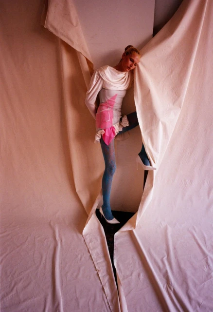 Fashion photo featuring a slender woman in a white dress and blue tights emerging from a tear in a large white cloth backdrop in a studio.