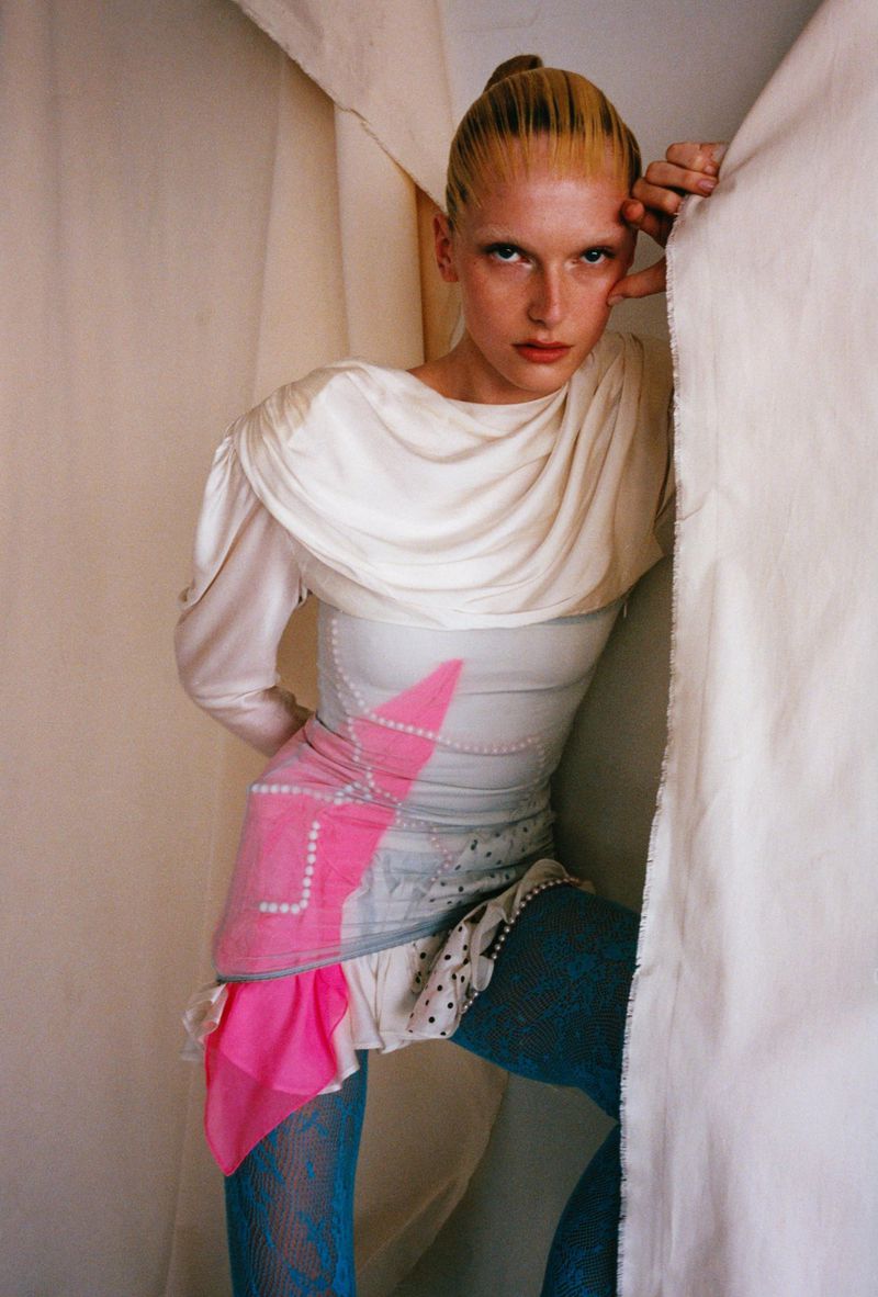 A fashion photo shows a slender adult woman with blond hair in a draped white top, patterned skirt, and blue patterned tights, posing indoors.