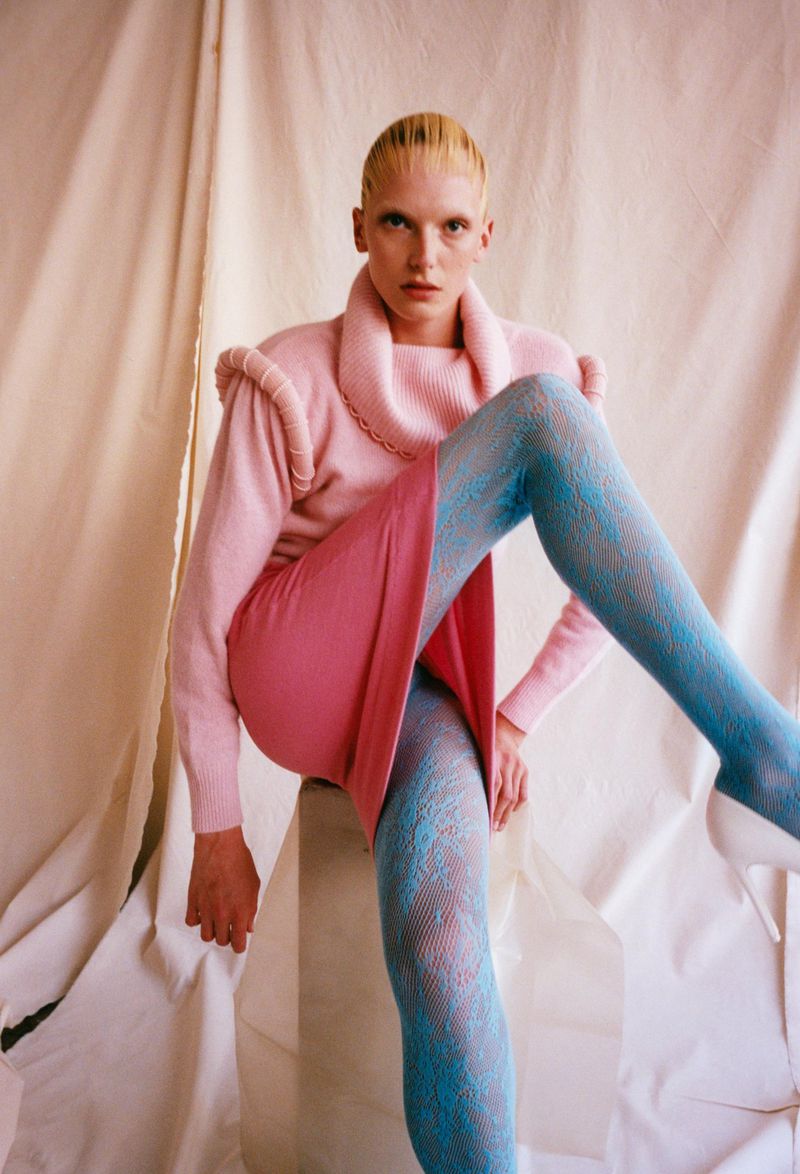 A female fashion model poses in a pink sweater and skirt with blue patterned tights in a studio with a draped backdrop.