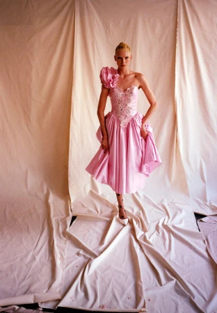A model poses in a one-shoulder pink dress with embellishments, flower detail, and white heels, against a draped off-white fabric backdrop.