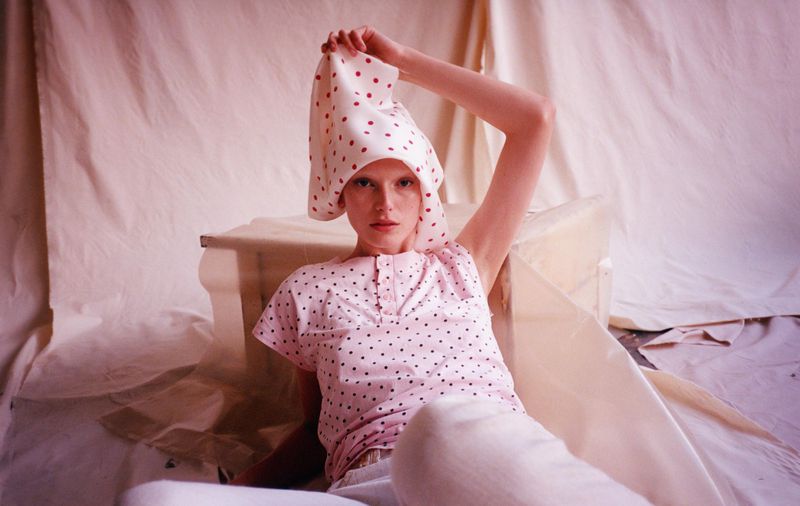 Young adult female in pink polka dot shirt and headscarf poses indoors against a draped fabric backdrop, looking directly at camera.