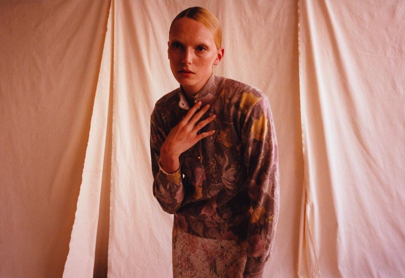 Adult female model with hand at neck, wearing floral-patterned sweater and skirt in front of a beige backdrop. Fashion, portrait, indoor.