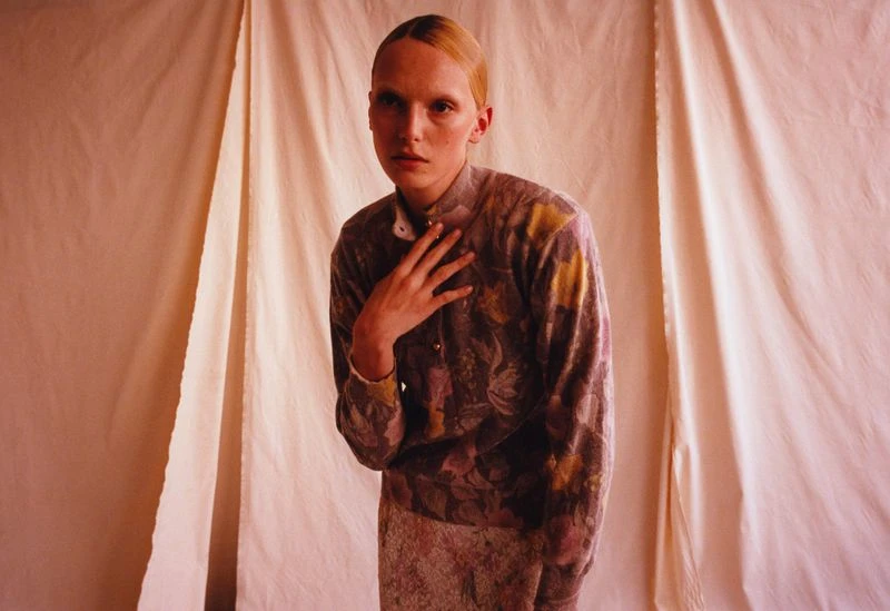 Adult female model with hand at neck, wearing floral-patterned sweater and skirt in front of a beige backdrop. Fashion, portrait, indoor.