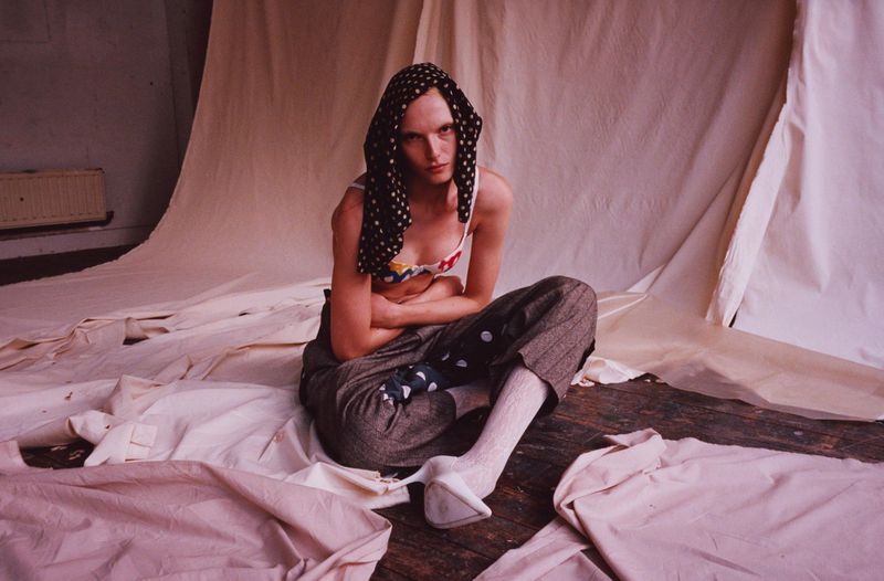 Non-binary person sitting in an indoor setting wearing a headscarf, patterned bra, and patterned trousers surrounded by white cloth.